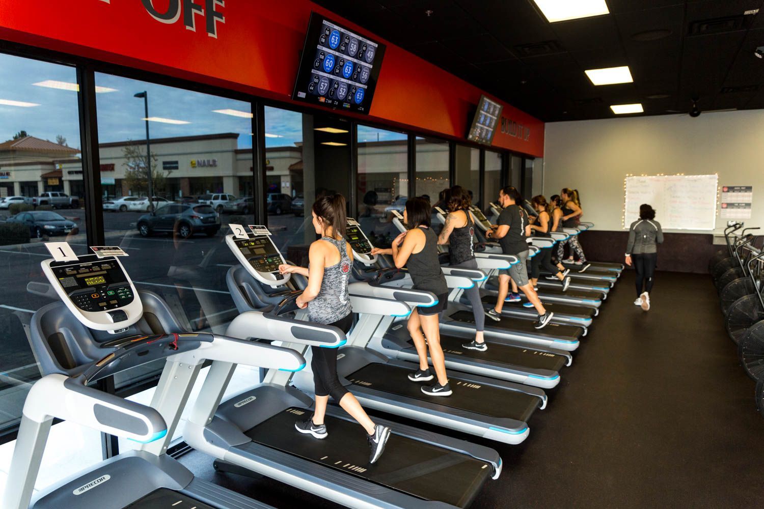 People running on treadmills in a brightly lit gym with windows overlooking a parking lot.
