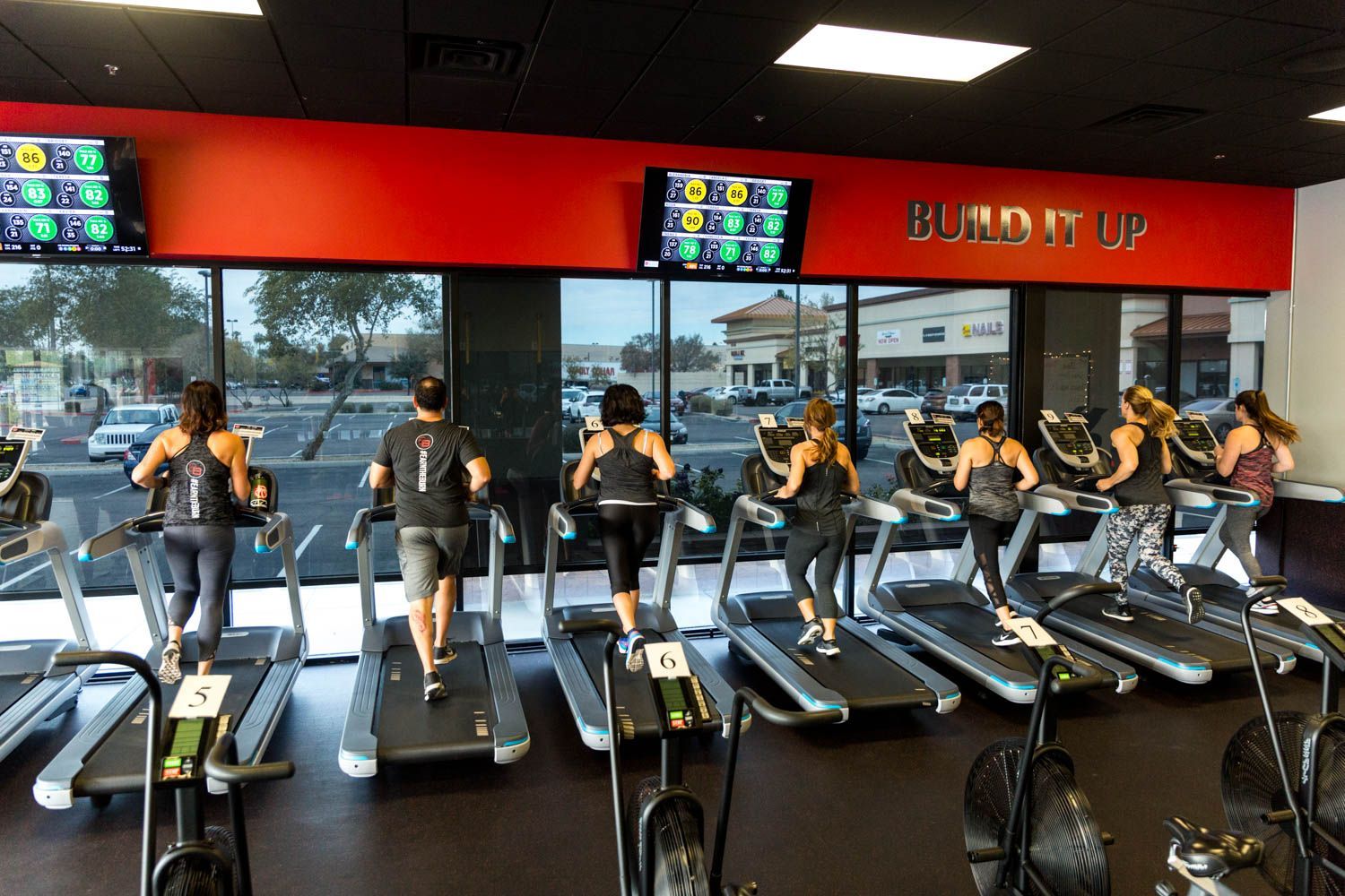 People running on treadmills in a brightly lit gym. The room has red walls and large windows with a cityscape view.