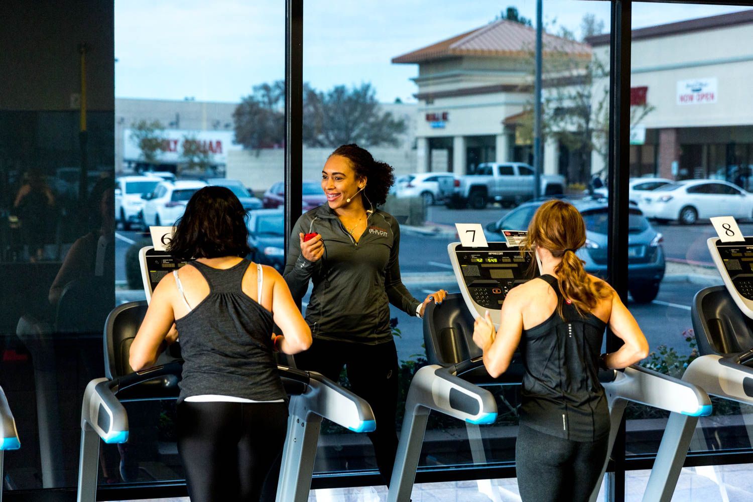 Three women exercising in a gym; two run on treadmills while the third speaks. The gym faces a parking lot.