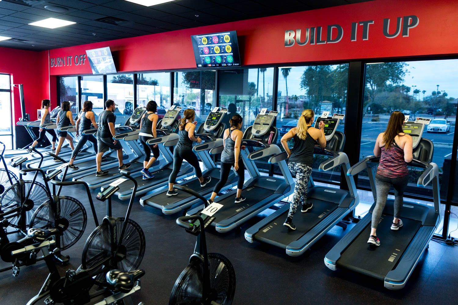 People running on treadmills in a brightly lit gym. Air bikes and large windows are also present.