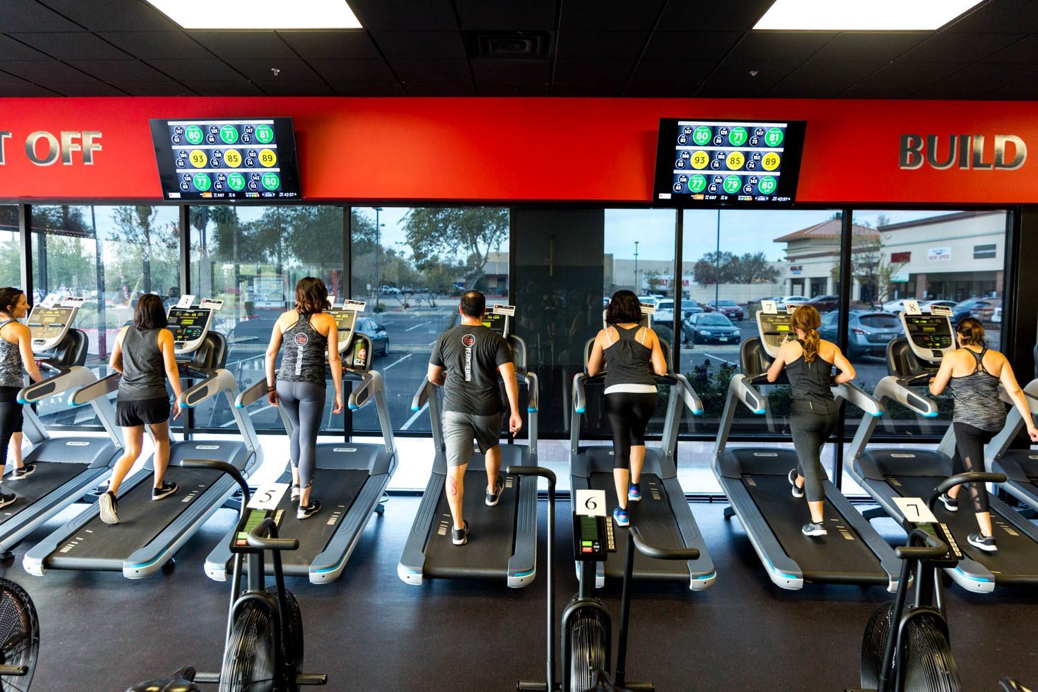 People on treadmills exercising in a gym with screens displaying data. Red and black color scheme.