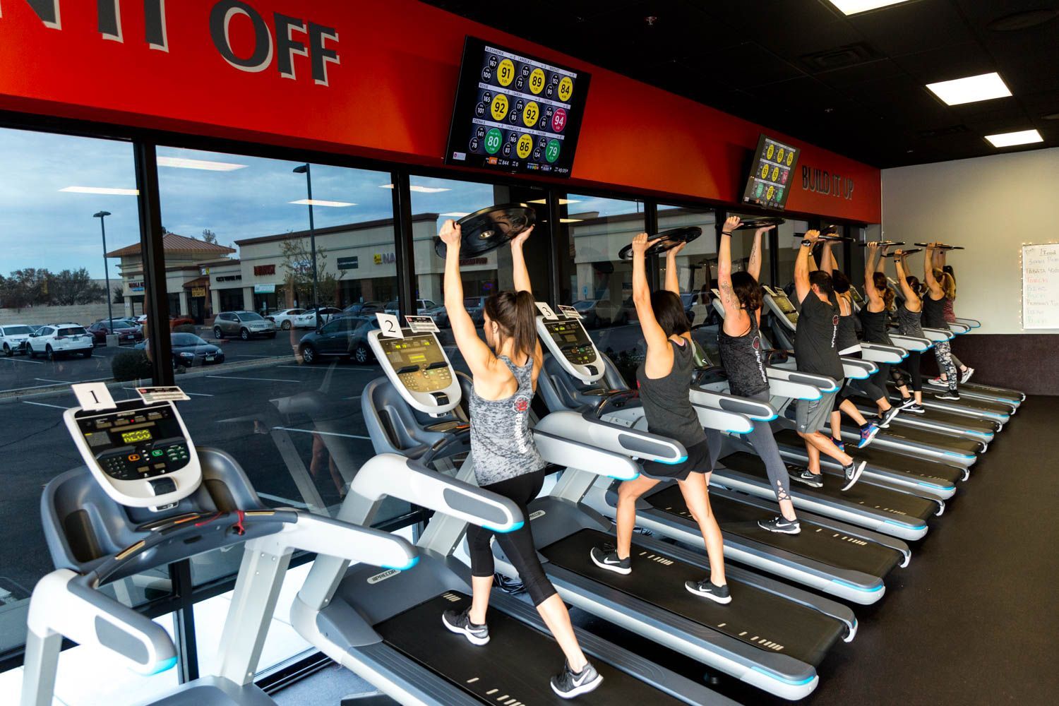 People exercising on treadmills, holding weights, in a brightly lit gym. Large windows overlook a parking lot.