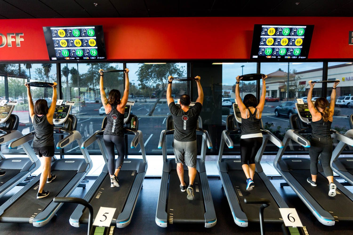 People on treadmills in a gym, holding weights above their heads.