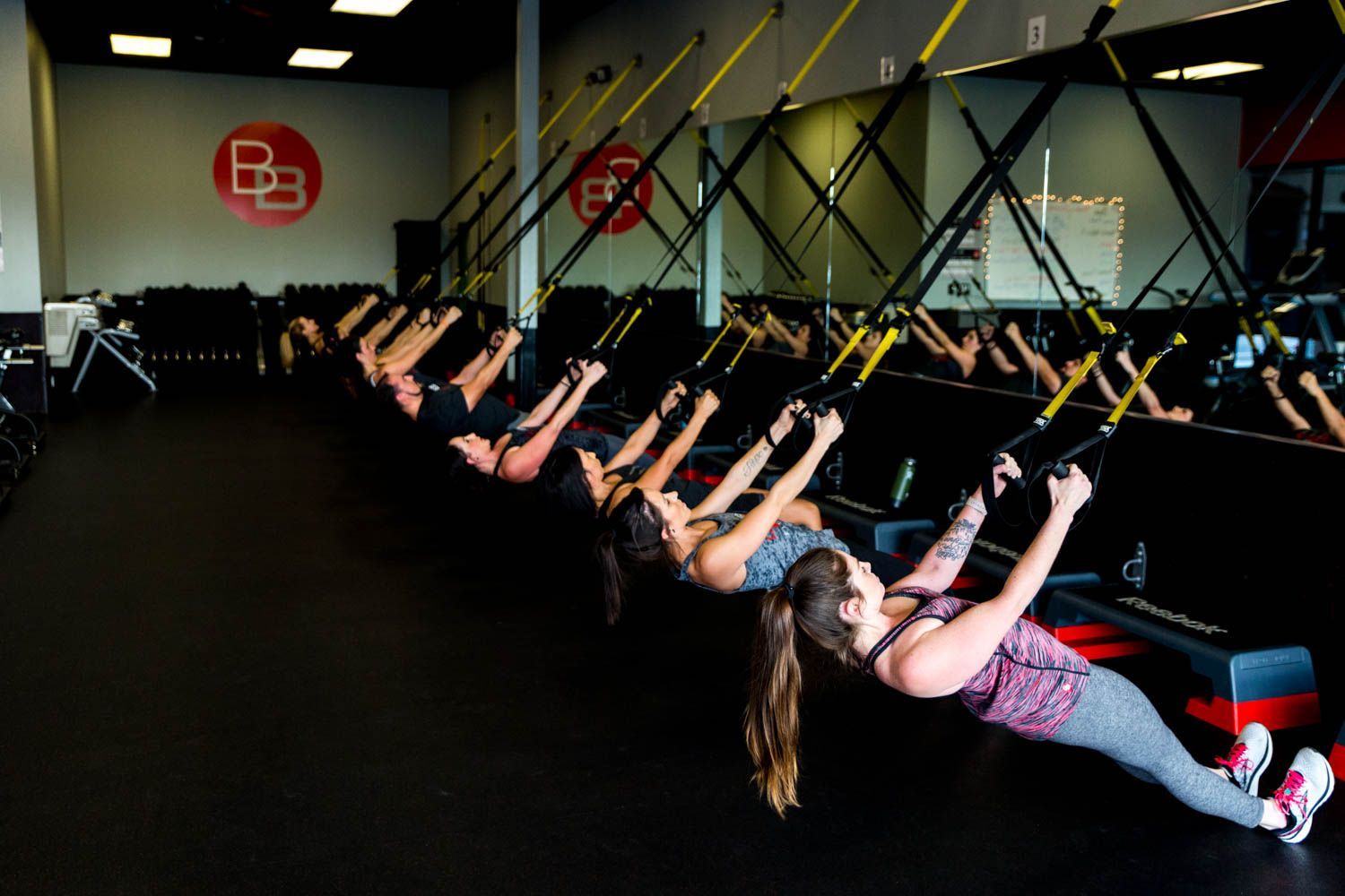 People in a gym use suspension trainers for exercise; they are leaning back, holding the handles.