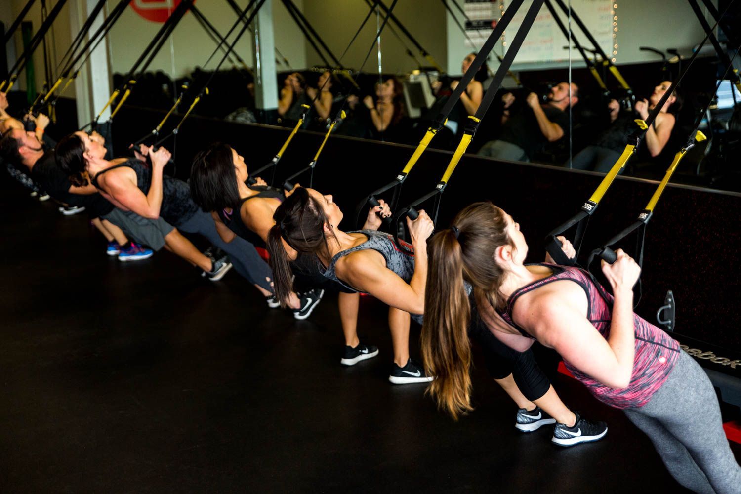 People exercising with suspension straps in a gym, leaning back, holding handles.
