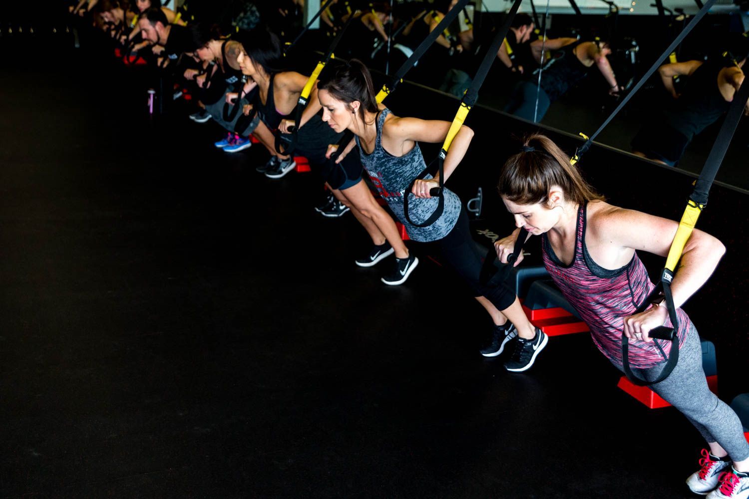 People in a fitness class doing suspended push-ups with black TRX straps. Dark room, black floor.