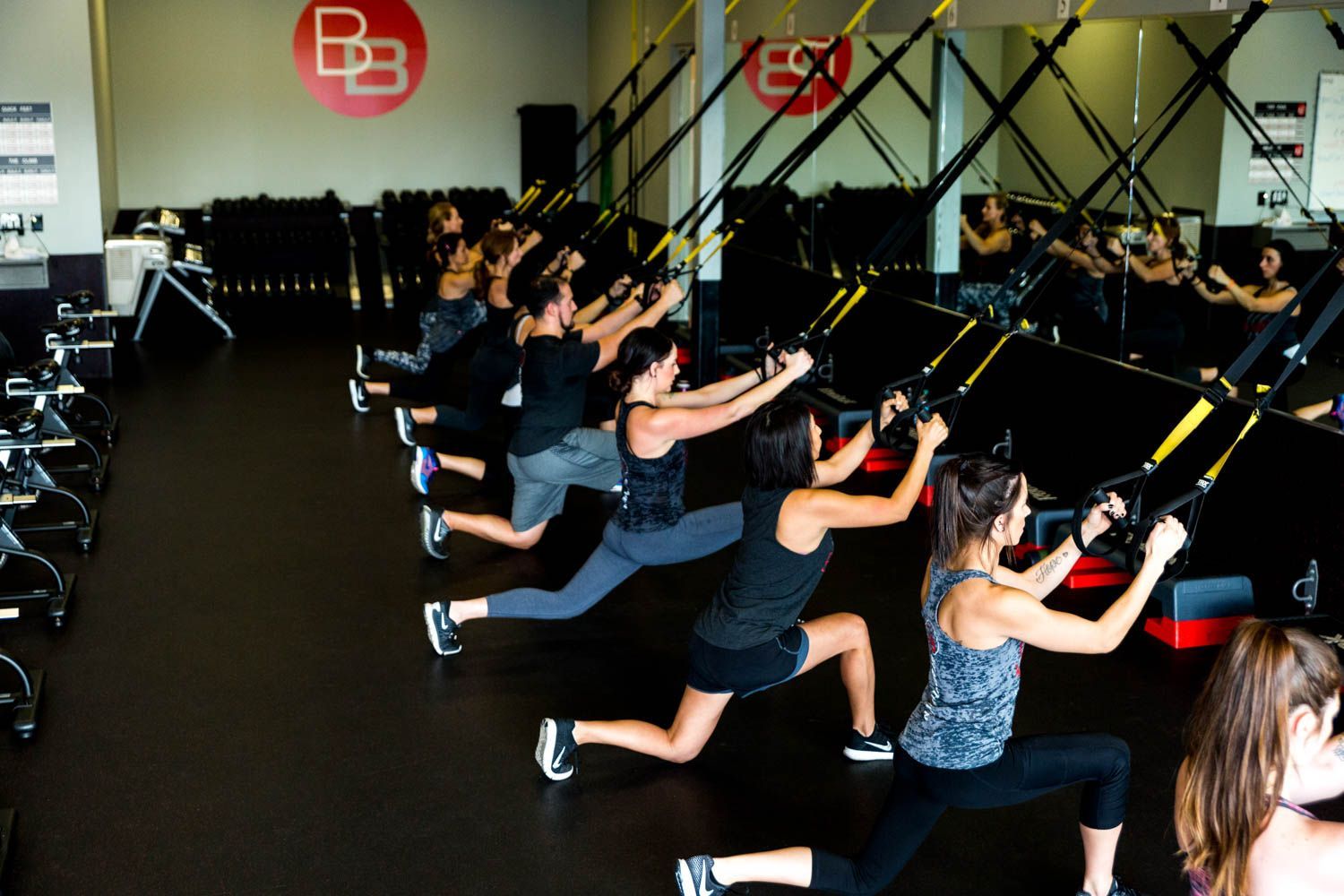People in a gym use suspension trainers for lunges; black floor, mirrored wall. 