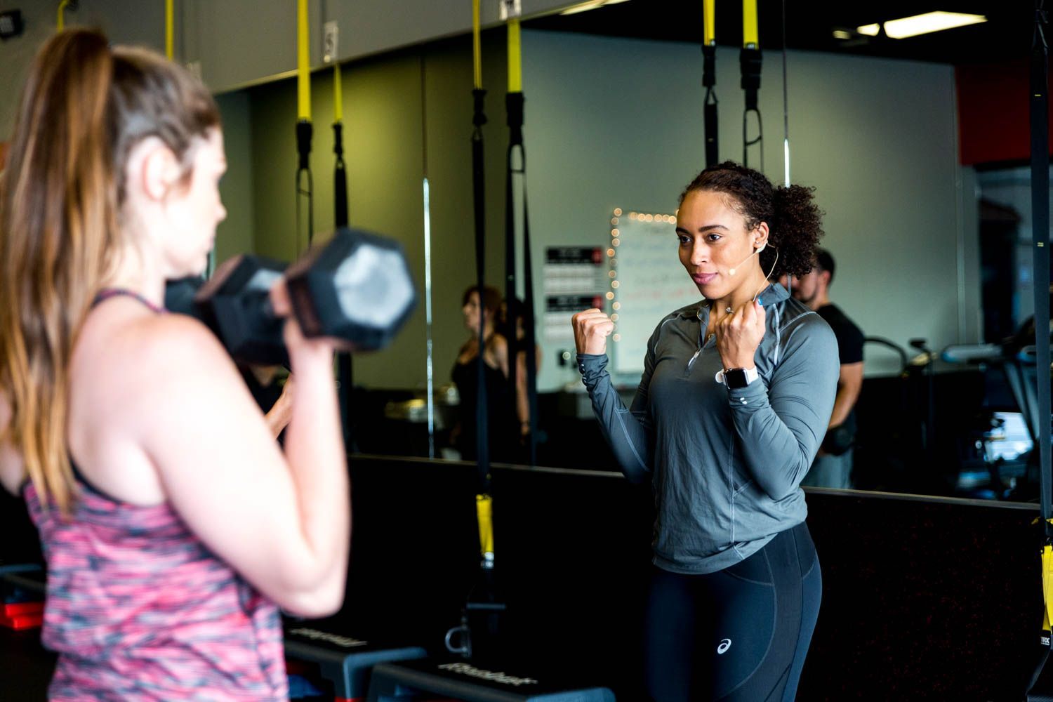 Woman lifting dumbbells, guided by instructor in gym.