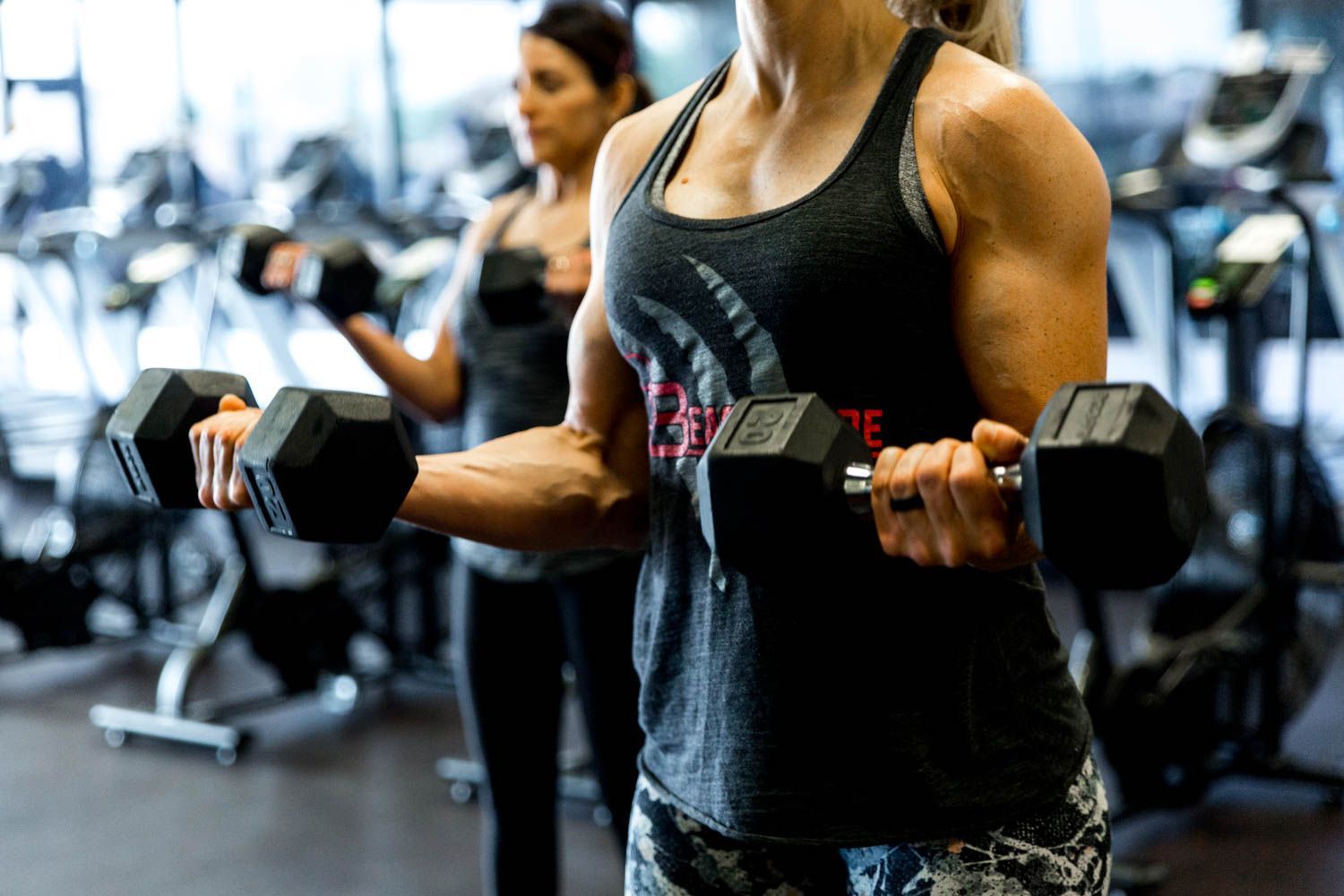 Two women lifting dumbbells in a gym, arms flexed. One woman in the foreground is wearing a gray tank top.