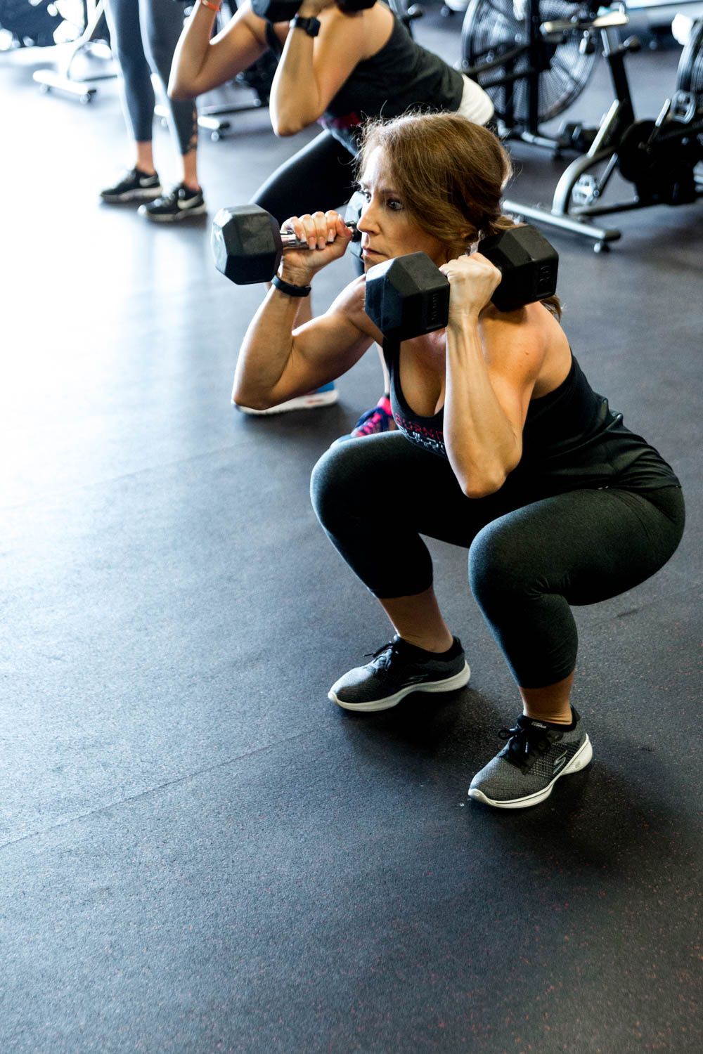 Woman doing dumbbell squats in a gym. She is wearing black workout clothes and holding dumbbells close to her chest.