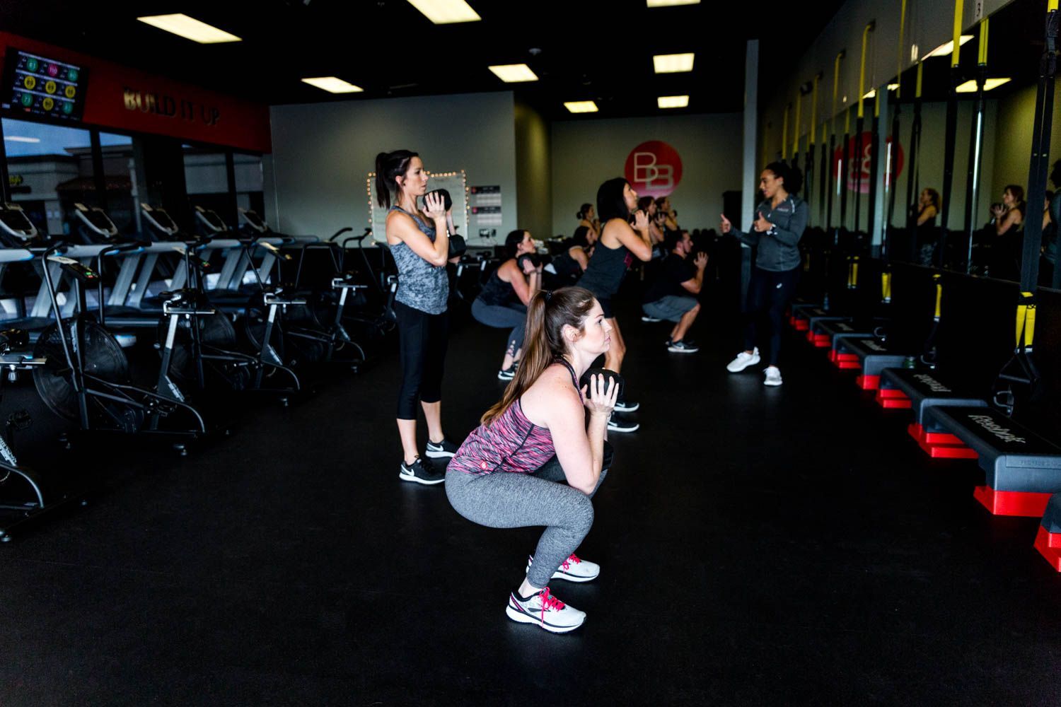 People doing squats with weights in a gym. A trainer watches, with exercise equipment.