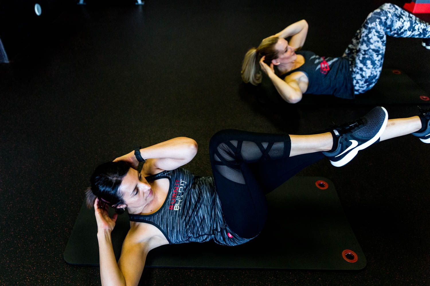 Two women on mats doing oblique crunches in a gym, black mats, gray tops, black leggings.