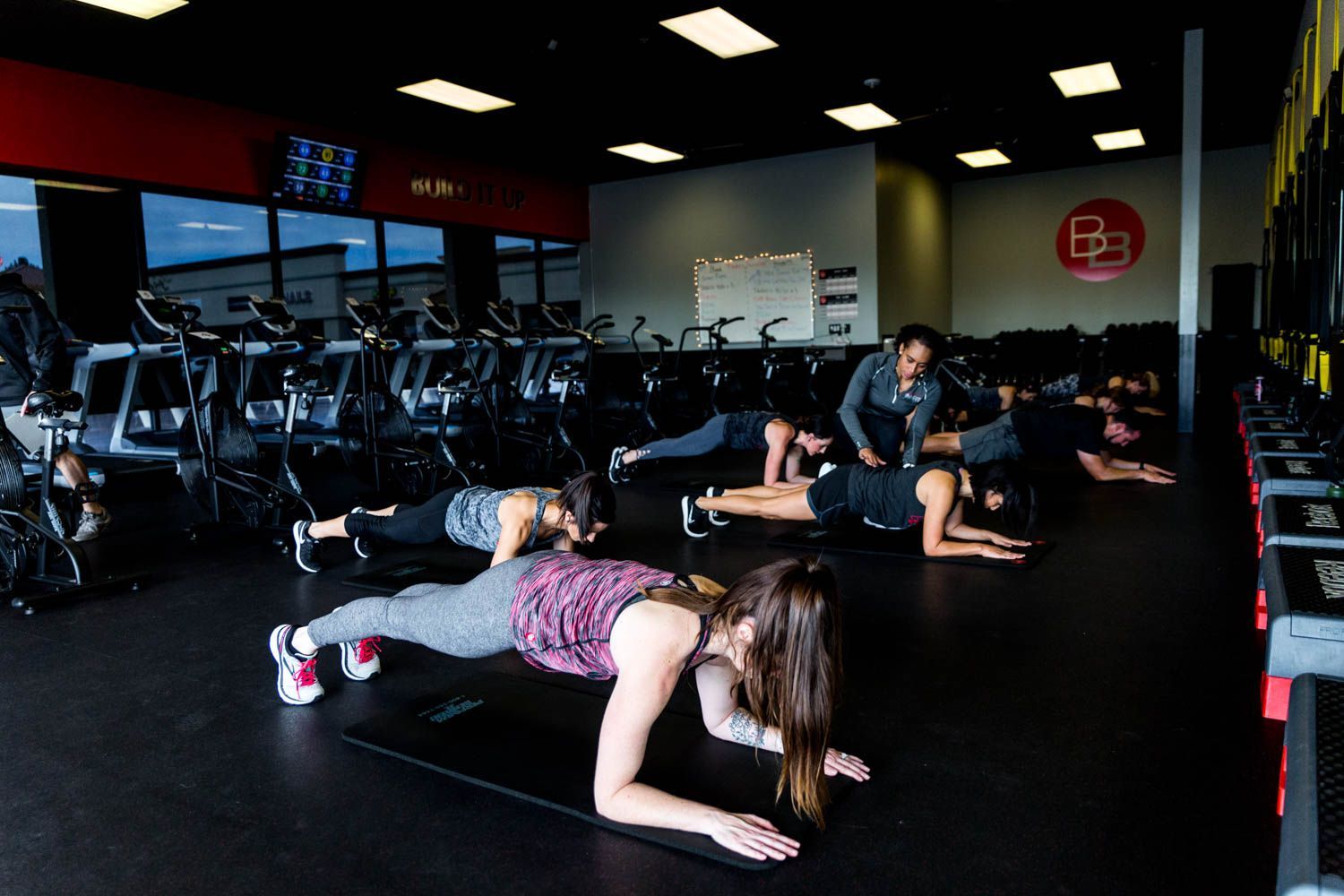 People doing planks on mats in a gym with stationary bikes in the background. Instructor assists.