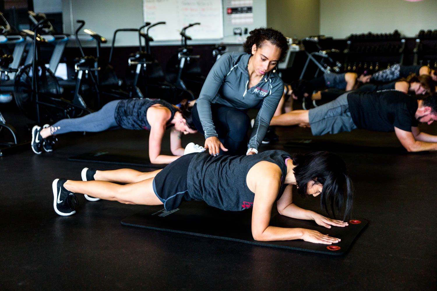 A fitness instructor demonstrating plank pose, correcting form of participant on mat in gym.