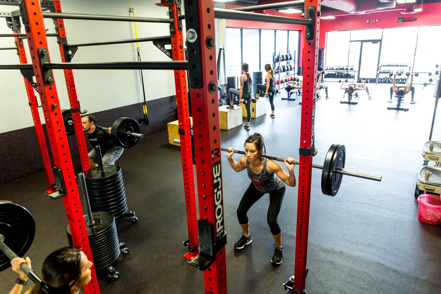 People lifting weights in a gym. A woman does a barbell squat, other people are in the background. Red and black equipment.