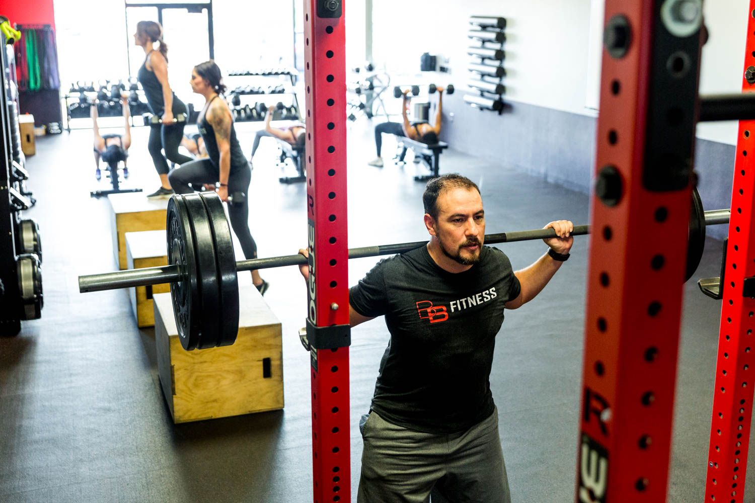 Man doing barbell squats in a gym, others doing various exercises in the background. Red weight rack.