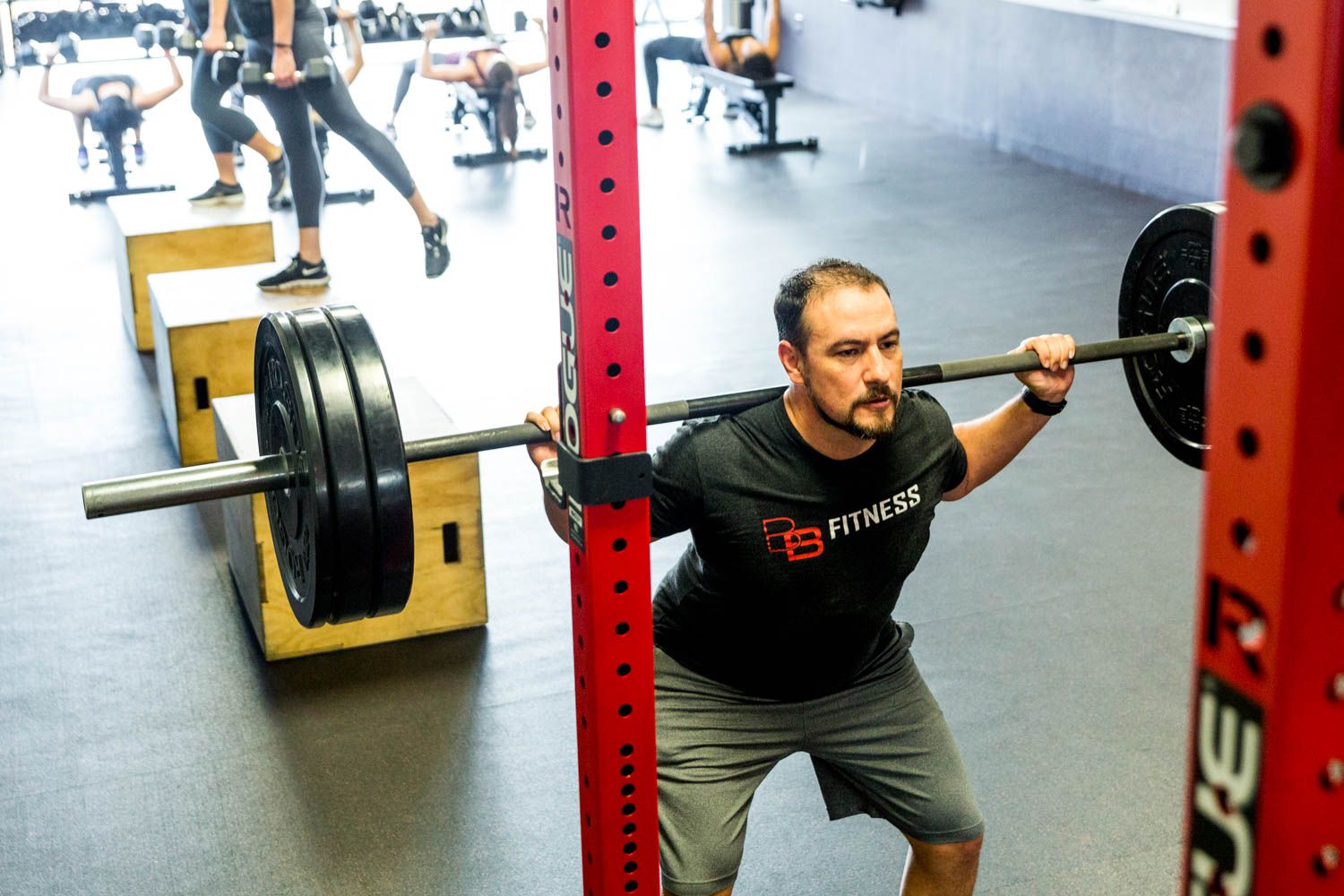 Man doing a barbell squat inside a gym with other people exercising in the background. Red weight rack in the foreground.