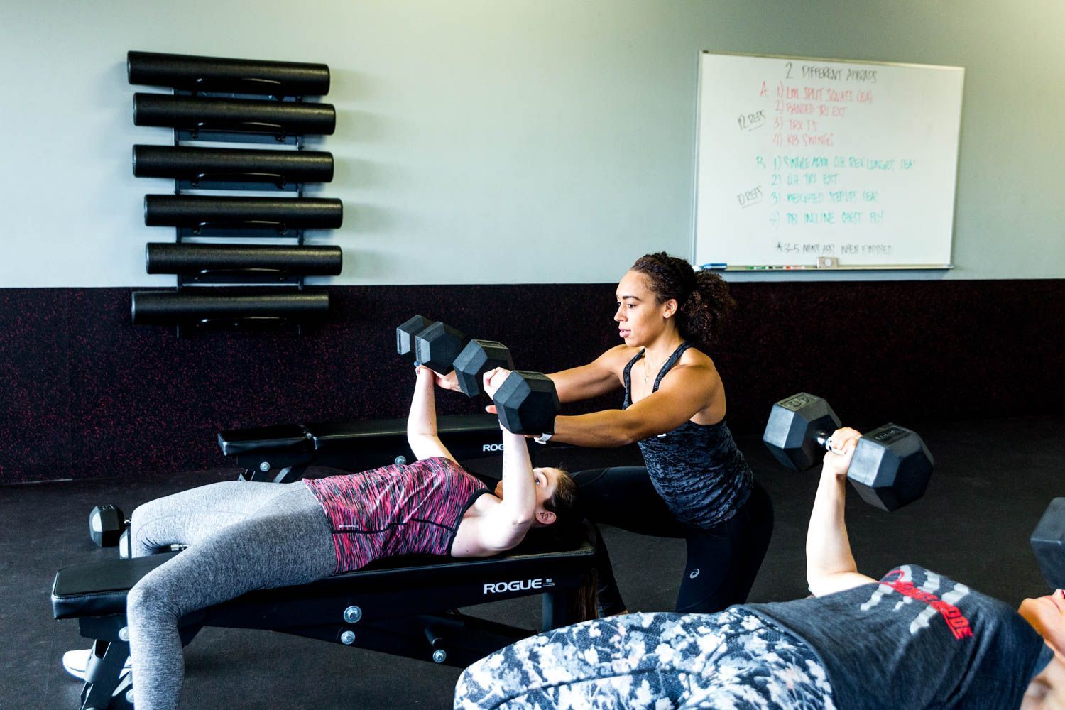 Woman spotting another woman doing dumbbell chest press on a bench in a gym.