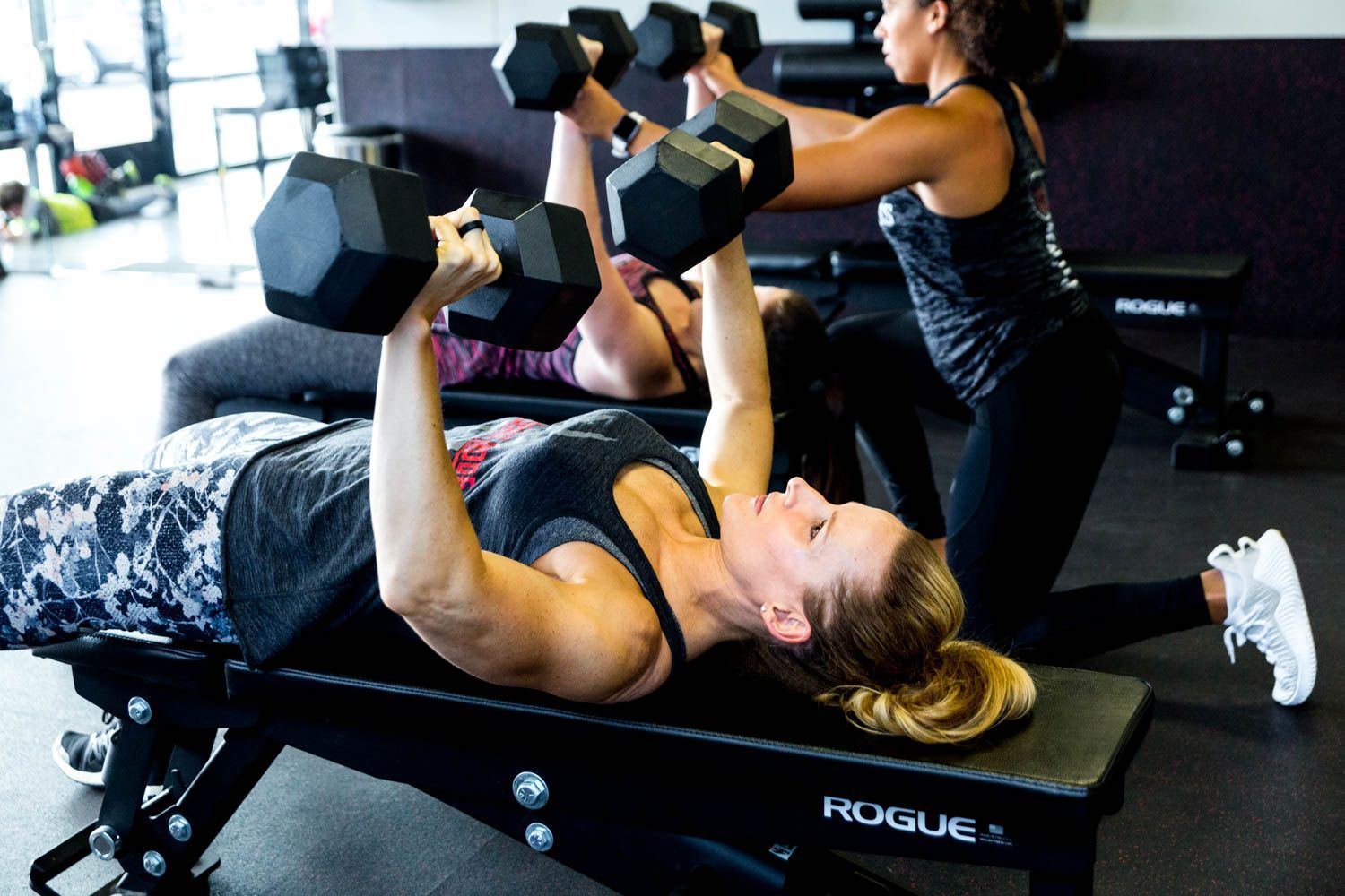 Two women lifting dumbbells while lying on a weight bench. One spotter assists. Gym setting.