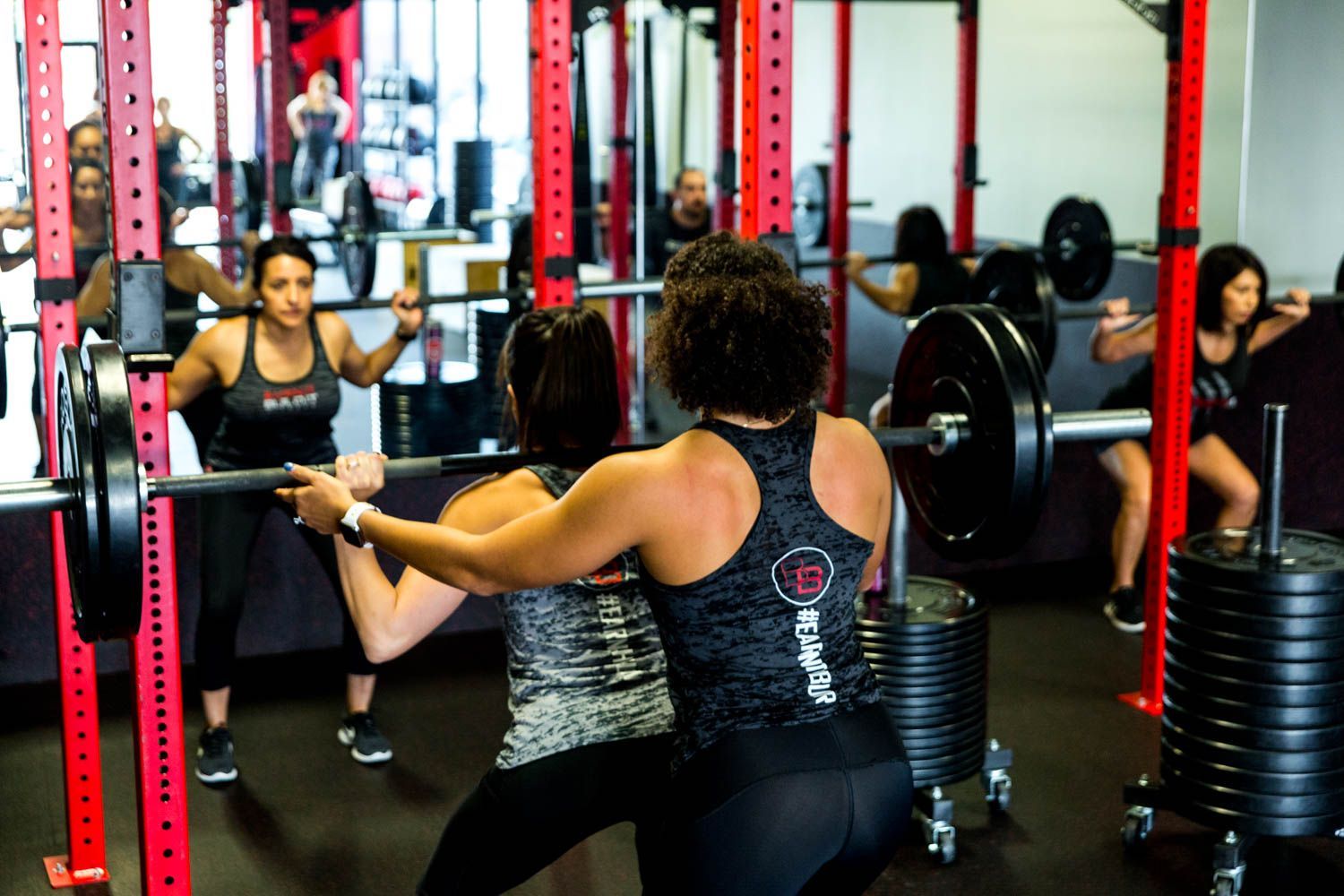 People squatting with barbells in a gym; a trainer assists one. Red weight racks.