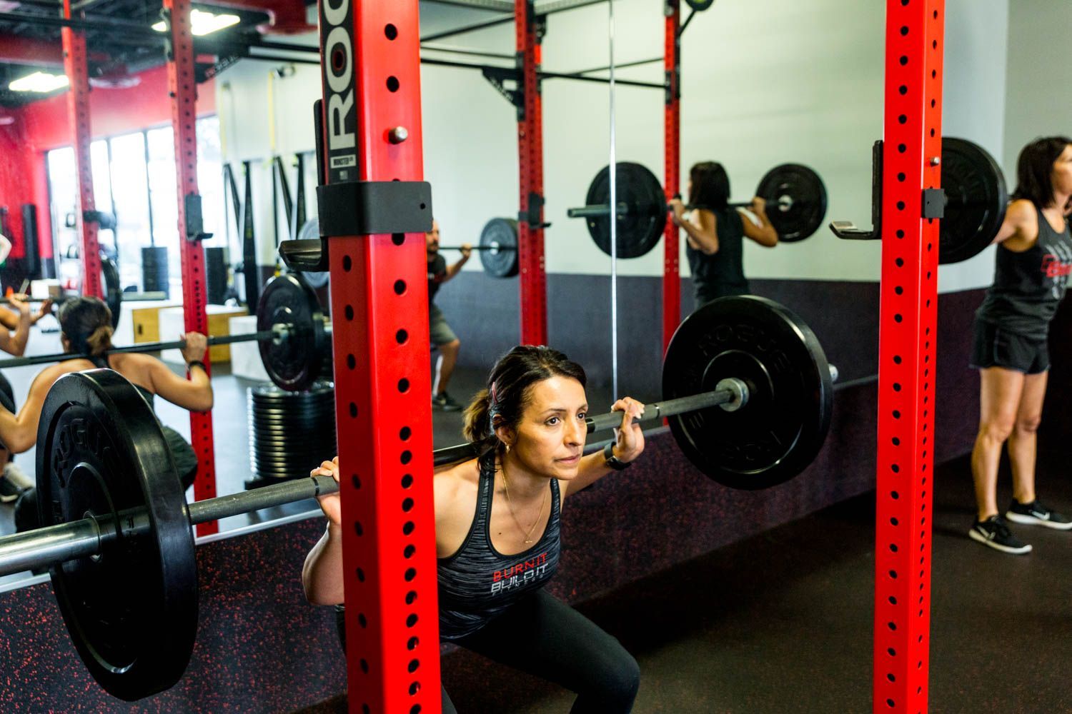 Woman squats with a barbell in a gym, red rack, others lifting in background.