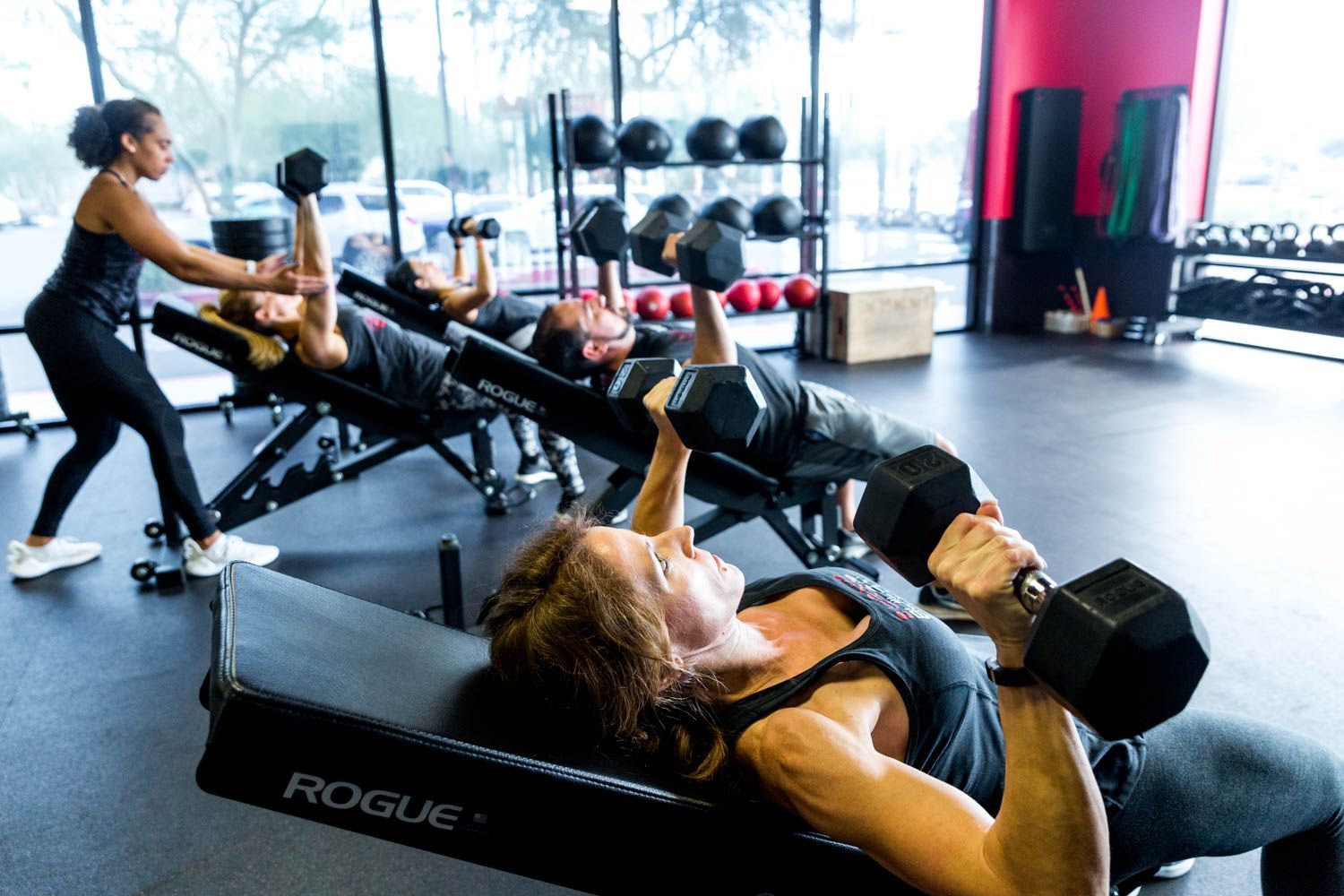 People lifting dumbbells on incline benches at a gym with assistance from an instructor.