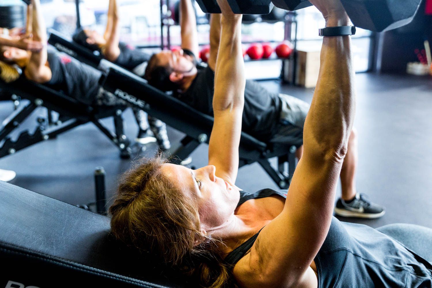 People in a gym on inclined benches, lifting dumbbells overhead.