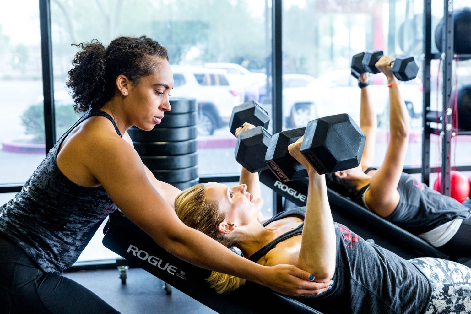 Trainer assisting woman lifting dumbbells on an incline bench in a gym.