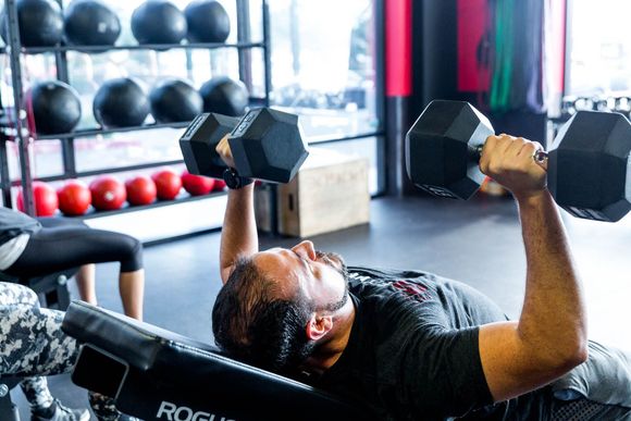 Man lying on a bench pressing dumbbells in a gym. Black dumbbells, red weights, and equipment are in the background.