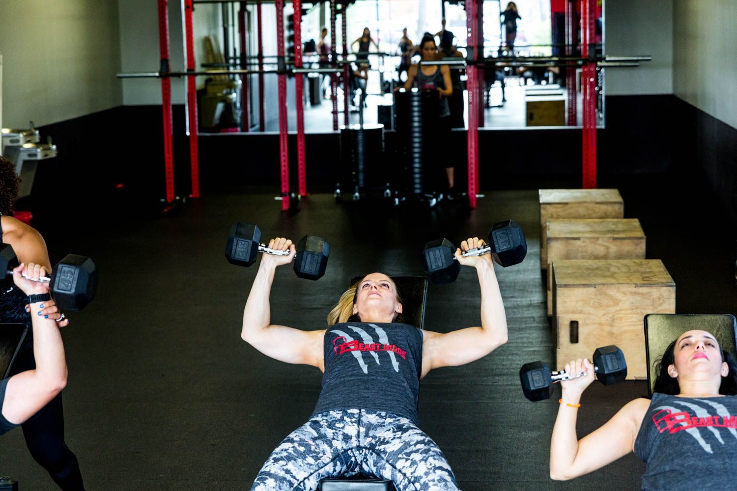 Three women in a gym, lying on benches lifting dumbbells. Dark flooring and red support beams.