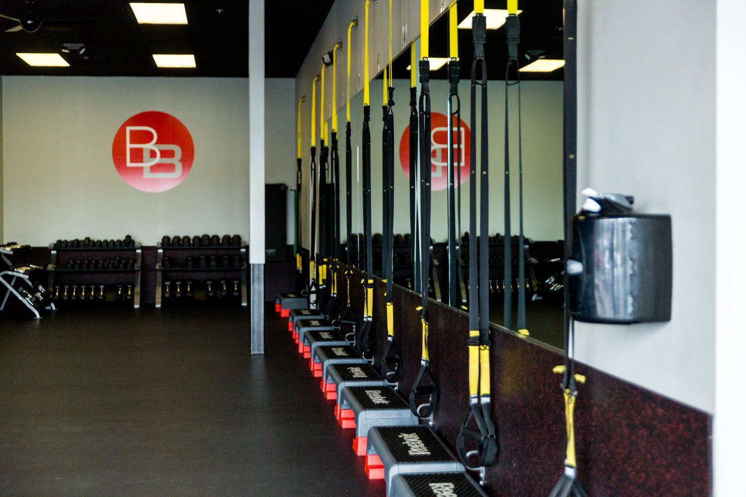 Gym interior with suspension trainers, weights, and a logo on the wall. Black floor, grey and white walls.