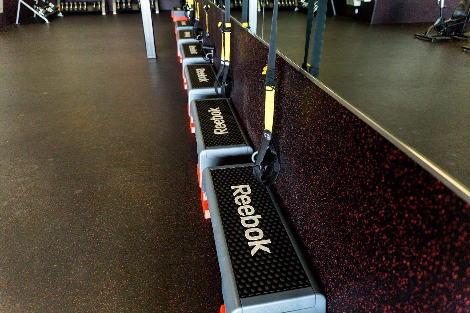 Row of Reebok step platforms in a gym, with black rubber flooring and a wall with suspension straps.