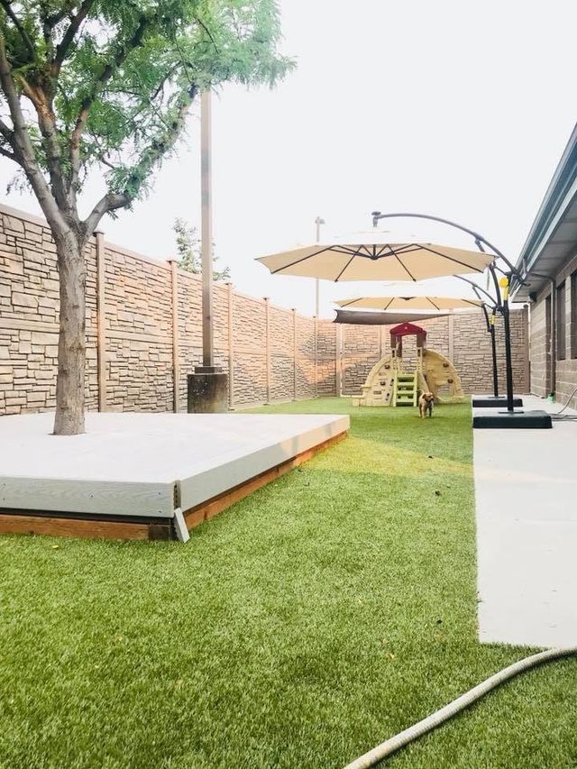 Outdoor patio with green turf, white platform seating, trees, and a shaded play area with a small slide.