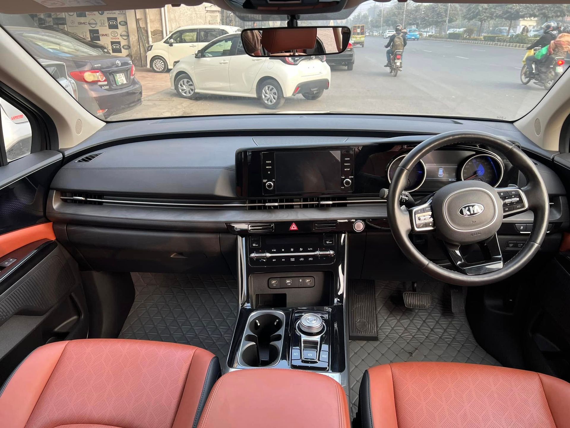 Interior view of a Kia Carnival. Dashboard is black with a touchscreen display, brown seats, and a black steering wheel.