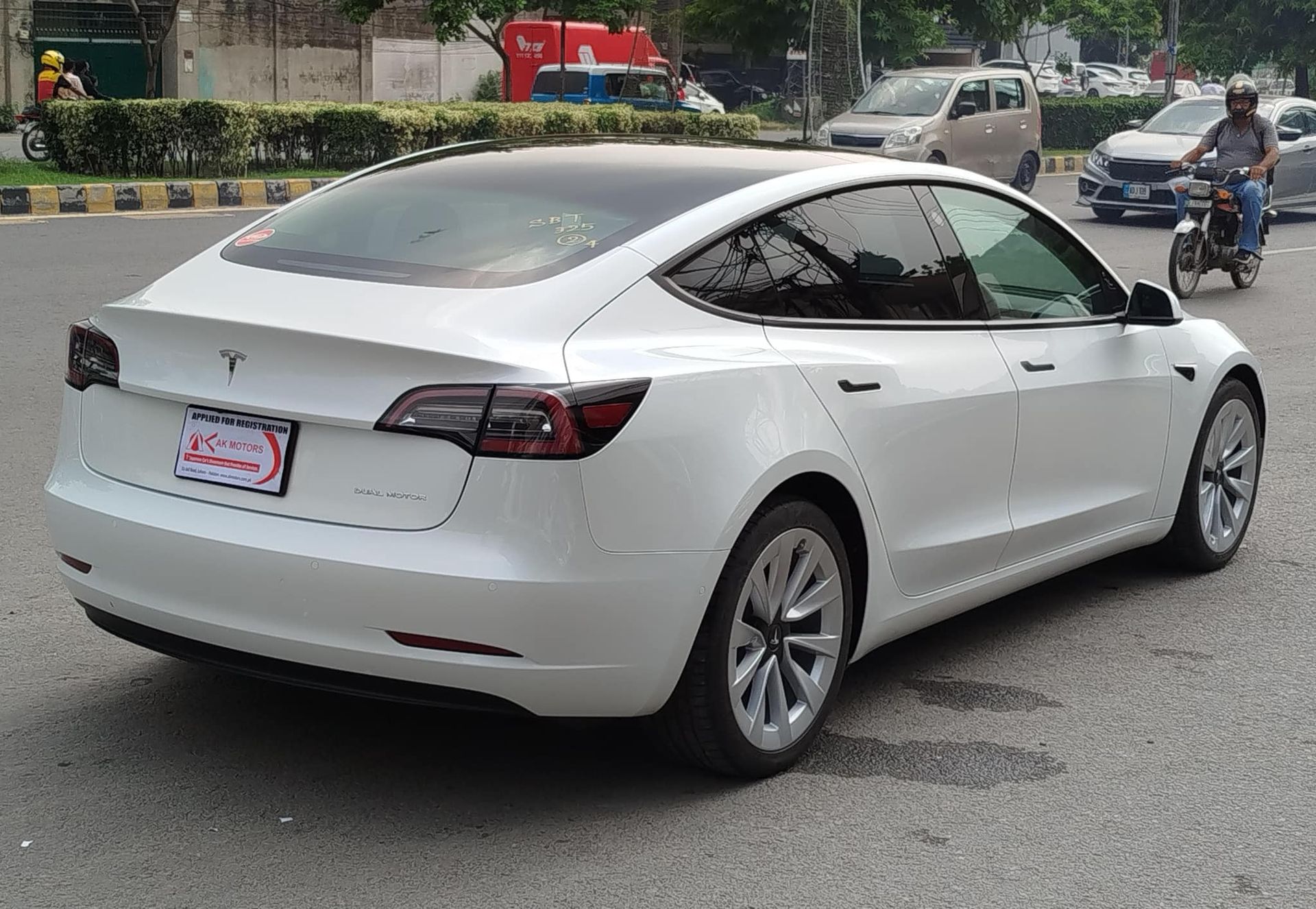 White Tesla car on a city street, rear view. Cars and a motorcycle are nearby.
