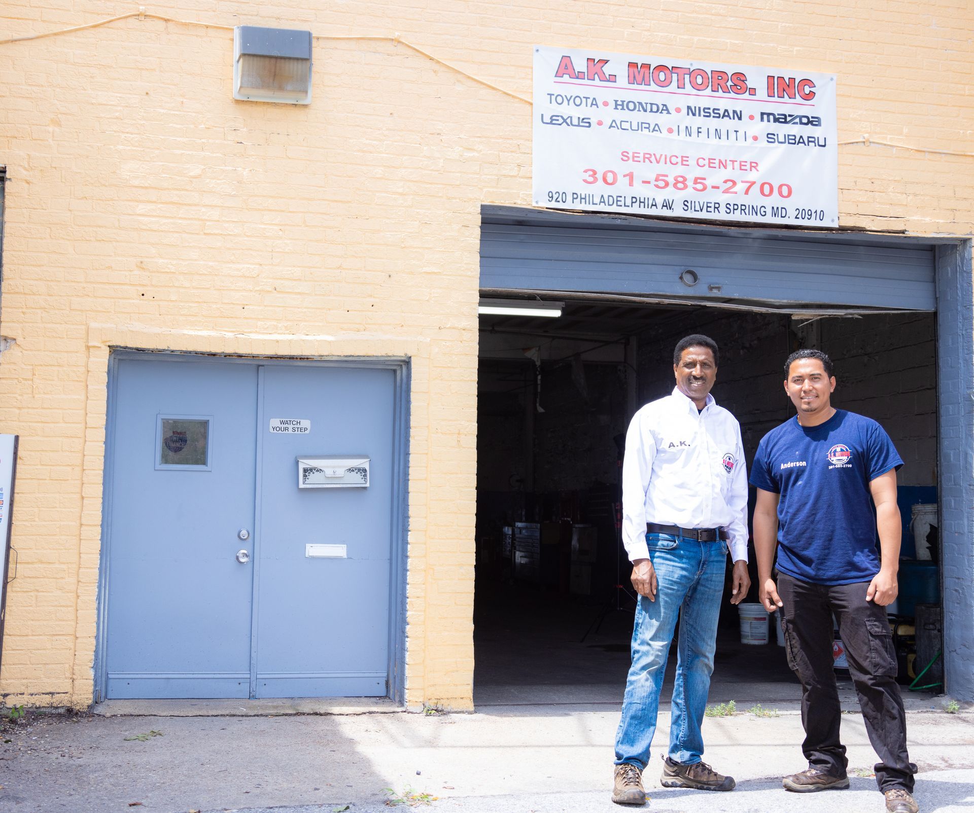 Two men standing in front of A.K. Motors, Inc. building. One in a white shirt and jeans, the other in a blue shirt and dark pants.