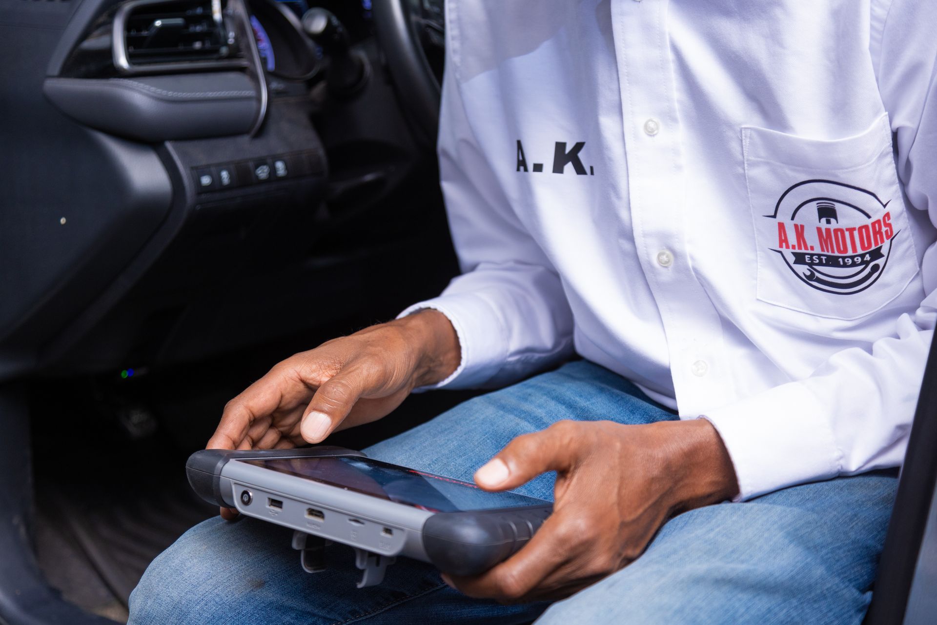 Person using a diagnostic tool inside a car; wearing a white shirt with a logo, and jeans.