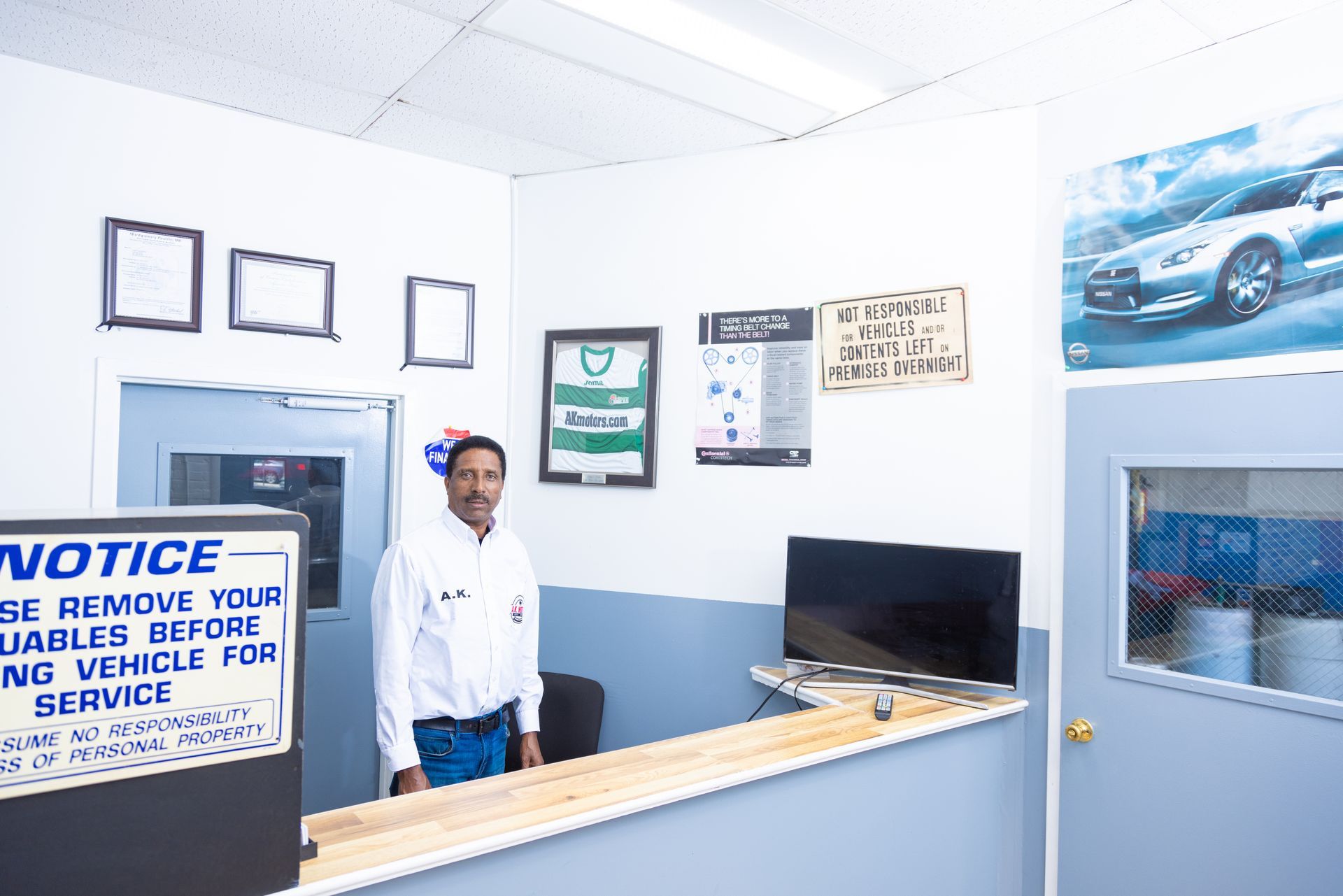 Man standing at a service counter in a car repair shop. White and blue walls, various signs, and a car poster are visible.