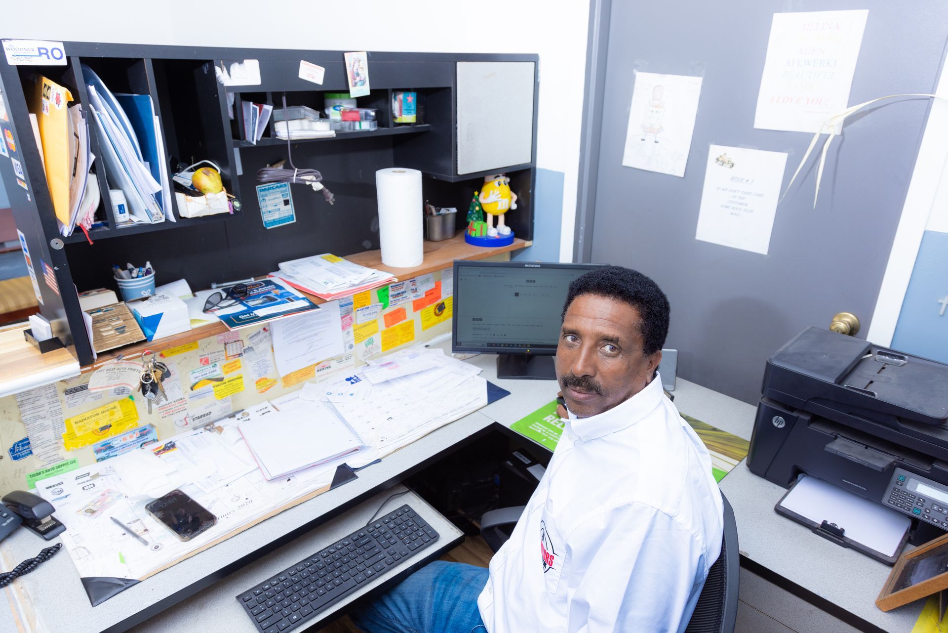 Man seated at a cluttered desk, looking at the camera. Office setting, computer, and printer visible.