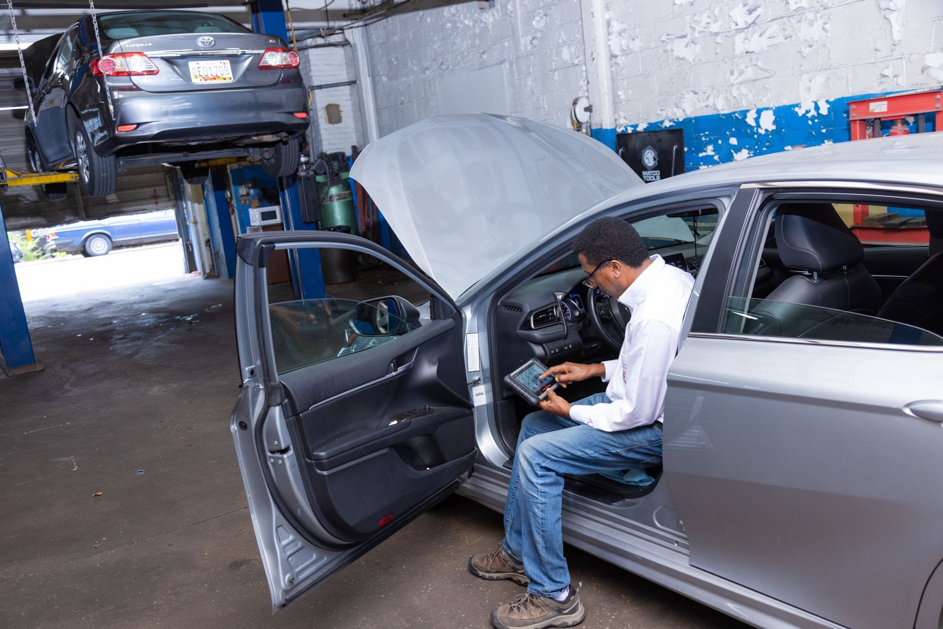 Mechanic using diagnostic tool in a car with open hood and door at a car repair shop. Another car is elevated.