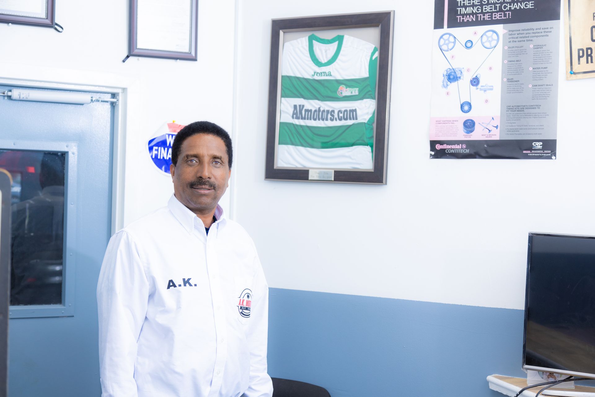 Man in white shirt stands in a room; a framed green and white jersey hangs on the wall.