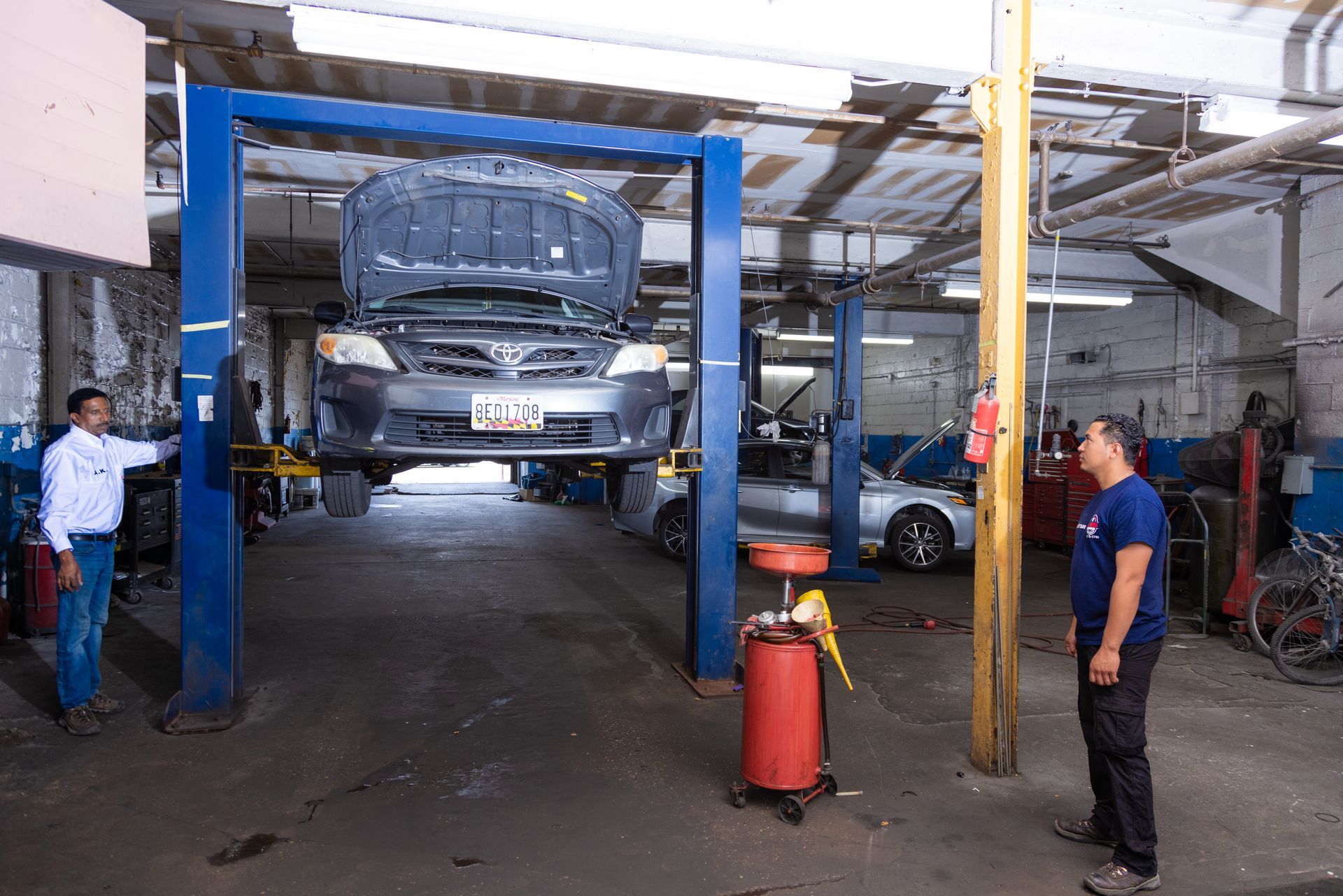 Car on a lift in a garage. Two men stand nearby; one points. Red oil container.