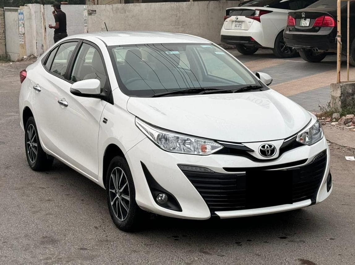 White Toyota Yaris sedan parked on pavement. Black and silver wheels, dark grille. Man standing in the background.