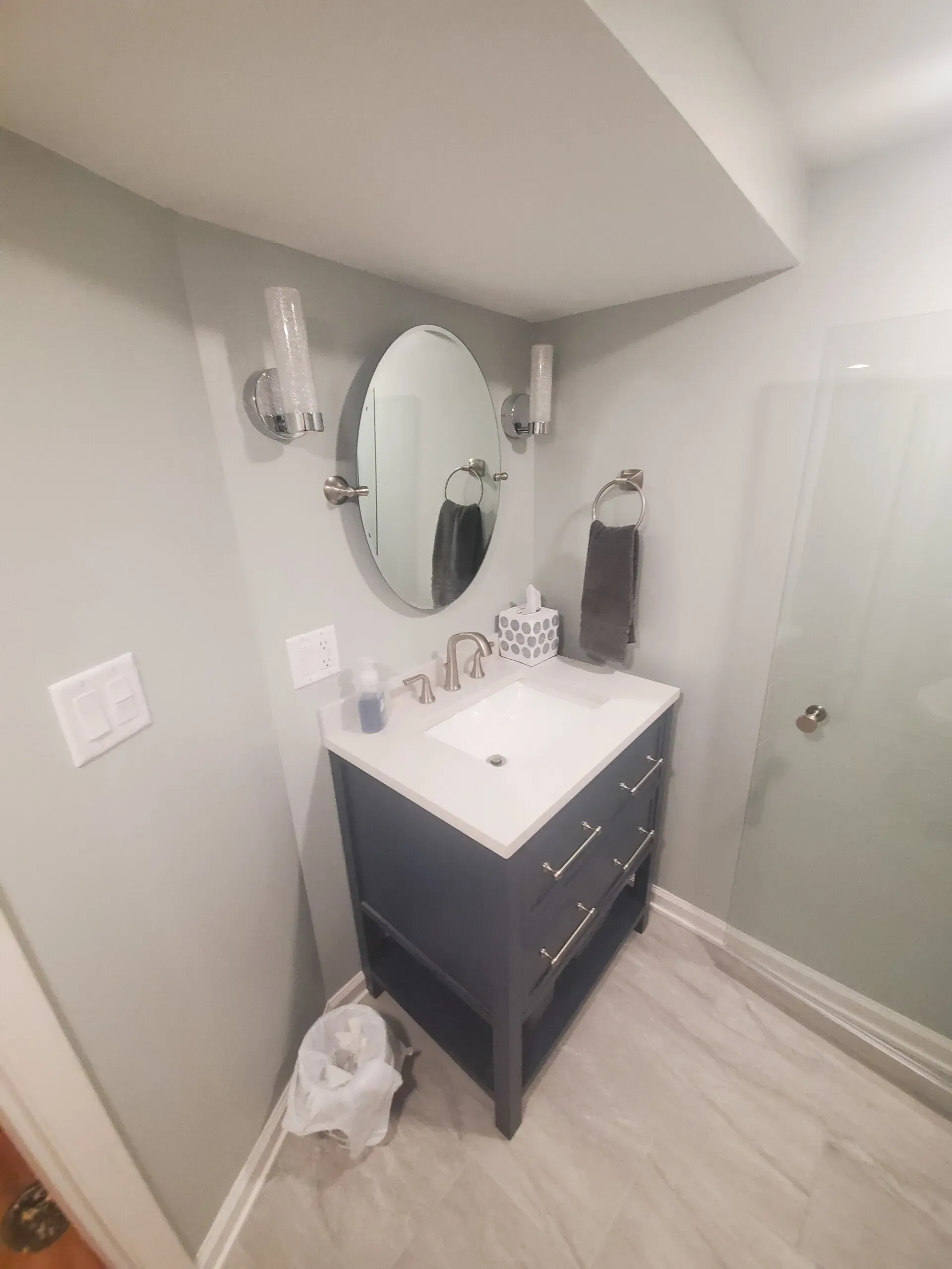 Small bathroom with blue vanity, oval mirror, sconces, and shower.