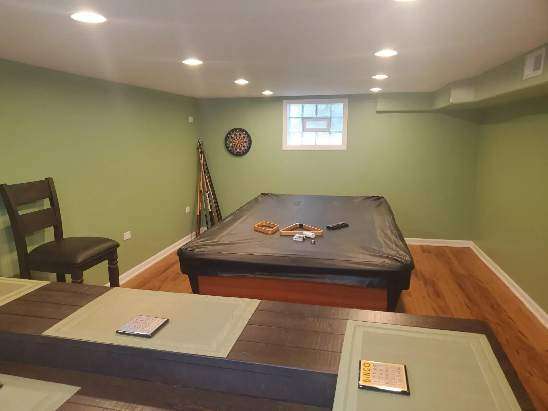 A basement rec room with a pool table covered in black, and a dining table.