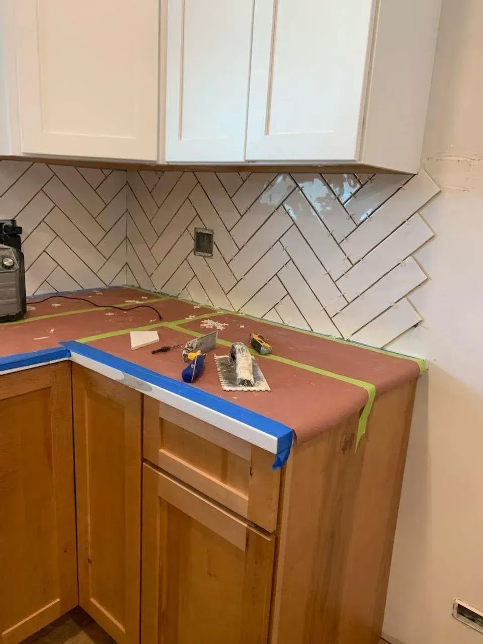 Kitchen corner with cabinets, countertop, and herringbone tile backsplash under construction.
