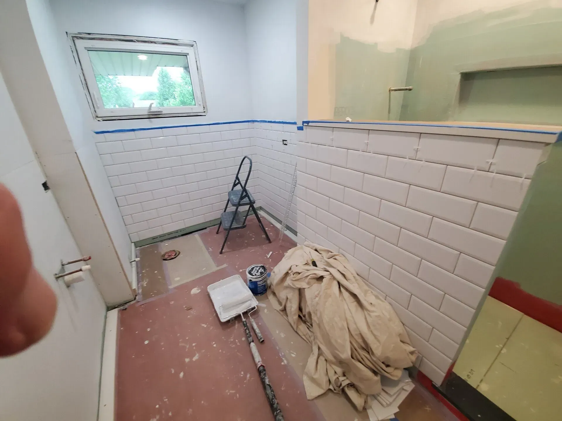Bathroom renovation in progress, walls tiled, ladder, paint supplies, and window.