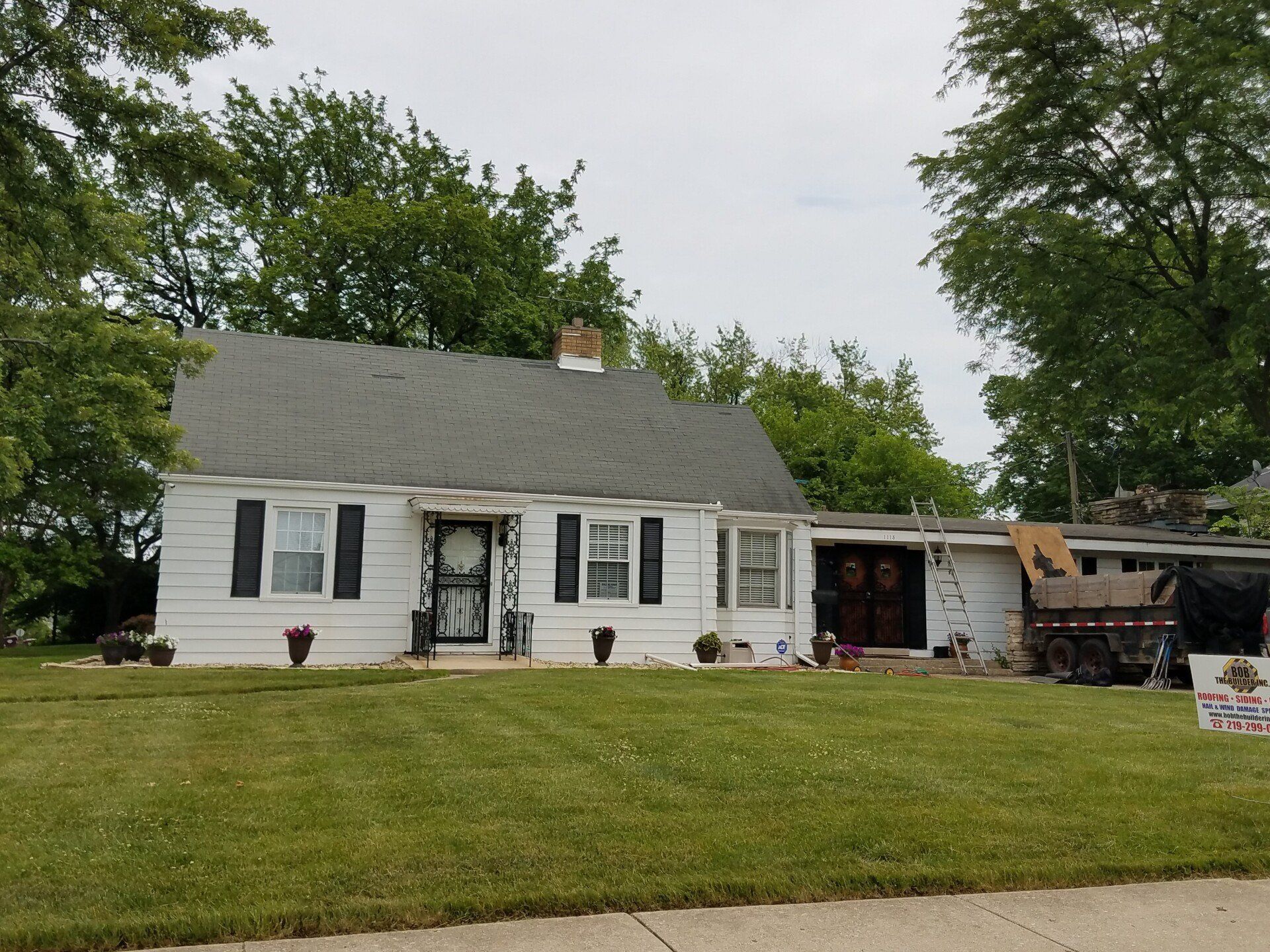White house with black shutters, green lawn, trees in the background, overcast sky.