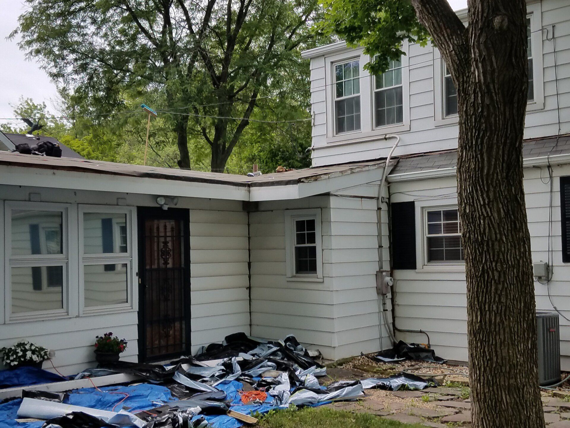 White house with roof damage and debris on the ground; trees in background.