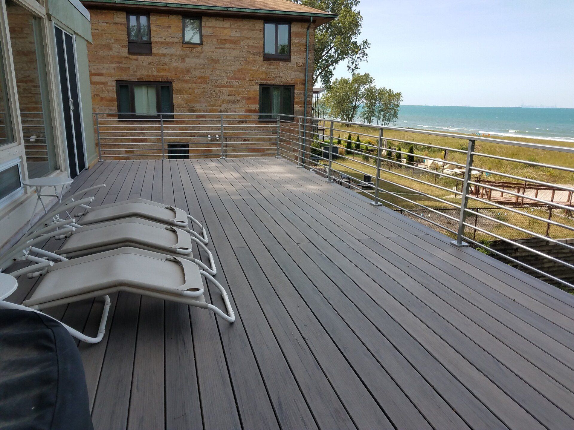 Deck with lounge chairs overlooking a beach and water, beside a brick building.